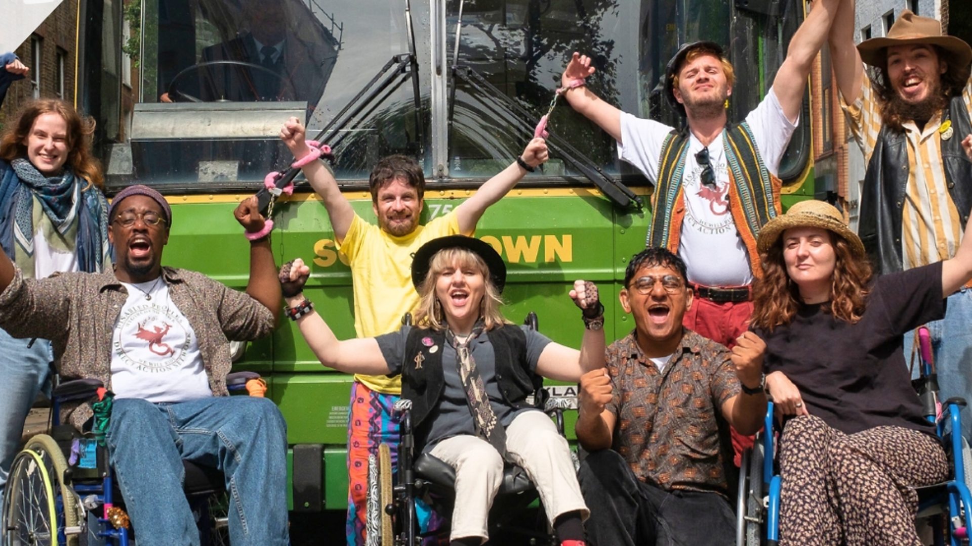A group of eight people wearing colourful clothes are cheering in front of a green bus. Some are standing while three people sit in wheelchairs. 