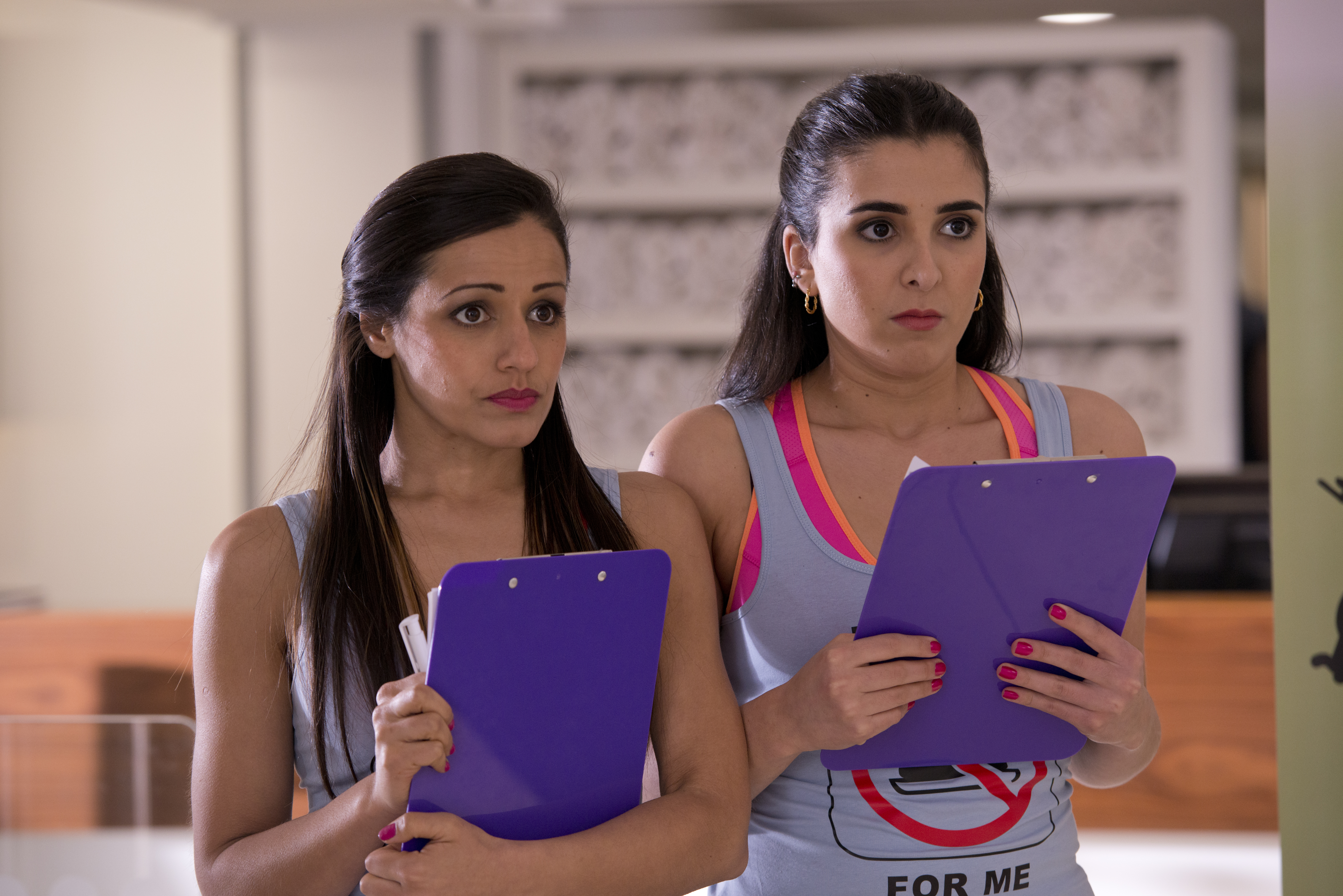 Production shot of two young women standing next to each other holding clipboards with anxious faces