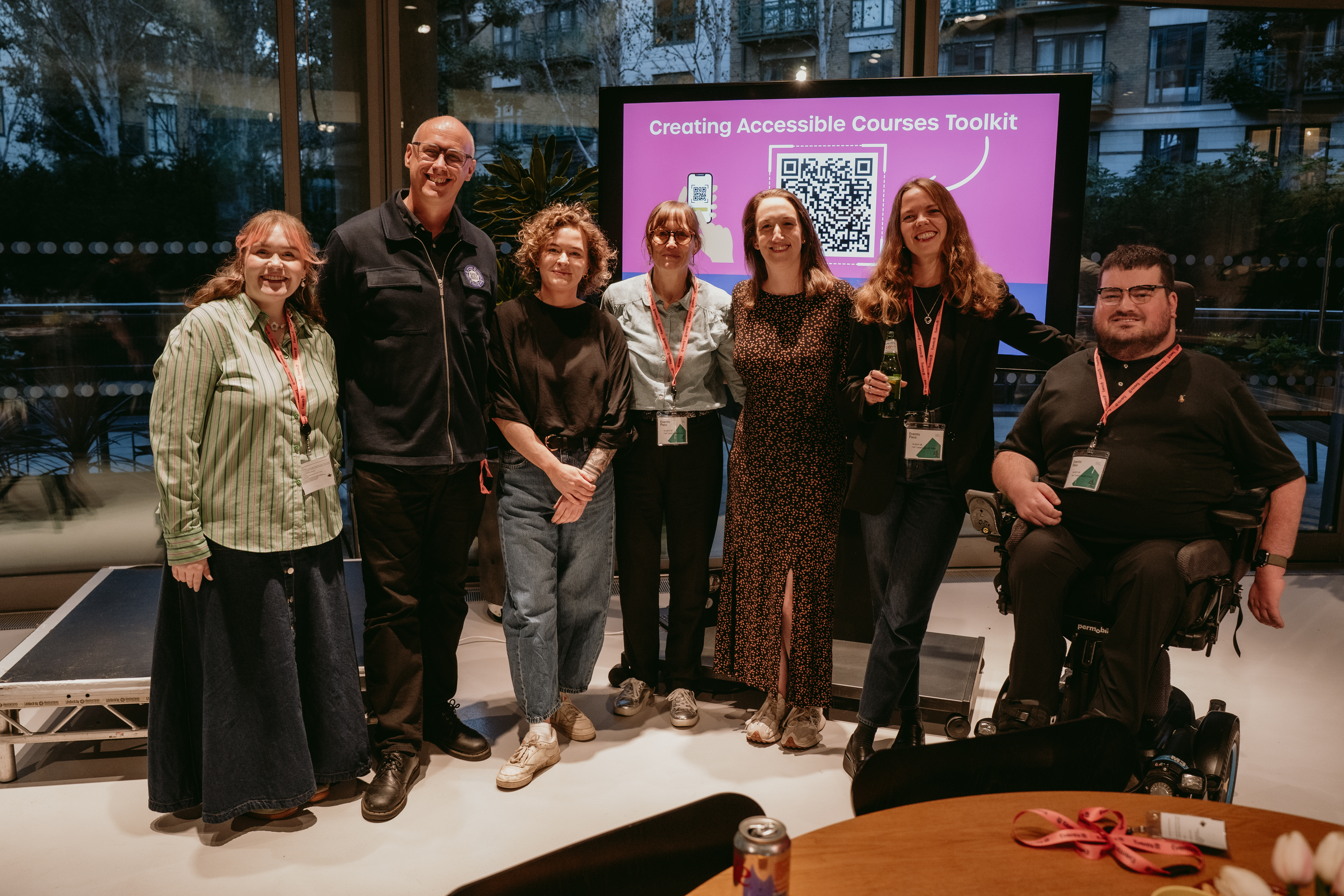 A group of people stand in a line smiling for a photo. Behind them a large TV screen, displaying promotion for the new Creating Accessible Courses Toolkit.