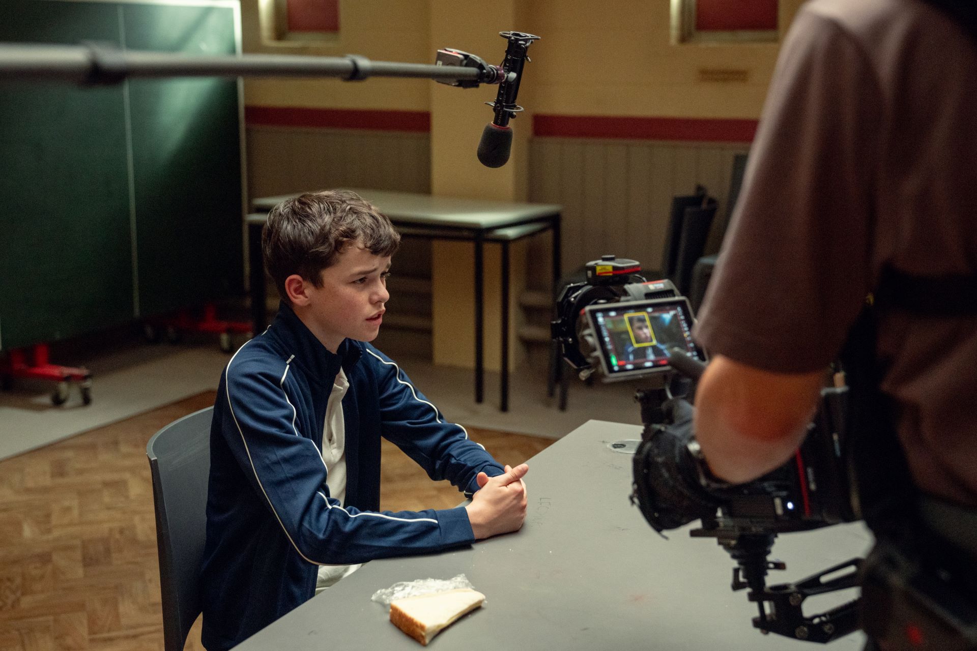 A BTS image of a production set with a boy sitting at a desk under a microphone and a camera