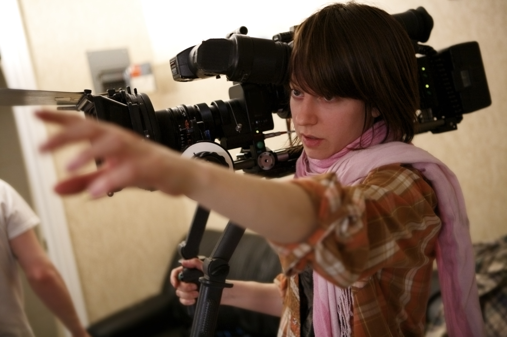 Close up of young person holding a large camera on their shoulder while reaching out with the other arm