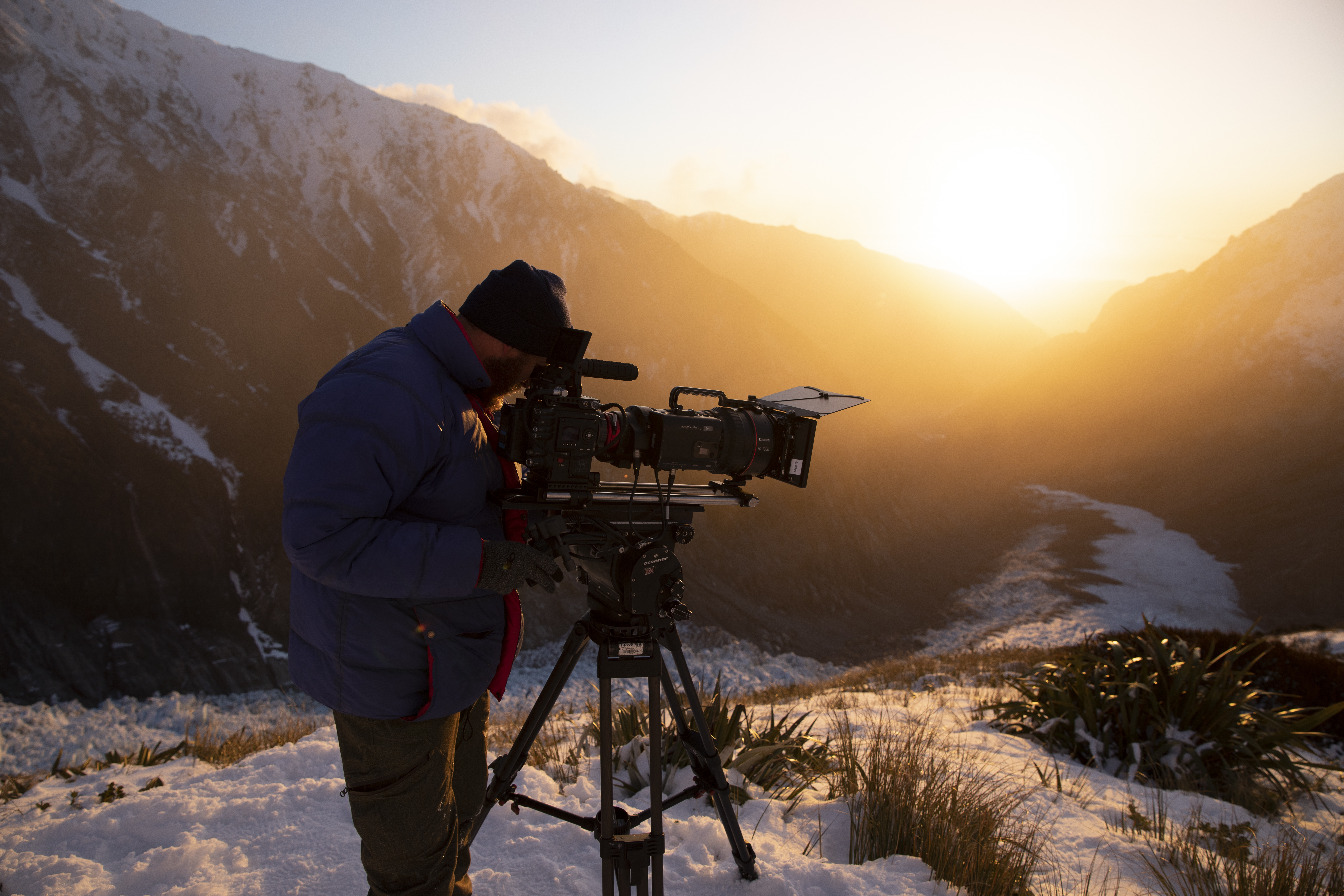 Camera operator is shooting a scene in the snow, wearing a heavy jacket and a hat.