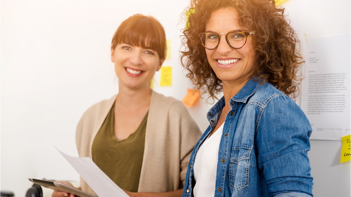Two women smiling at the camera holding a mobile device and a piece of paper