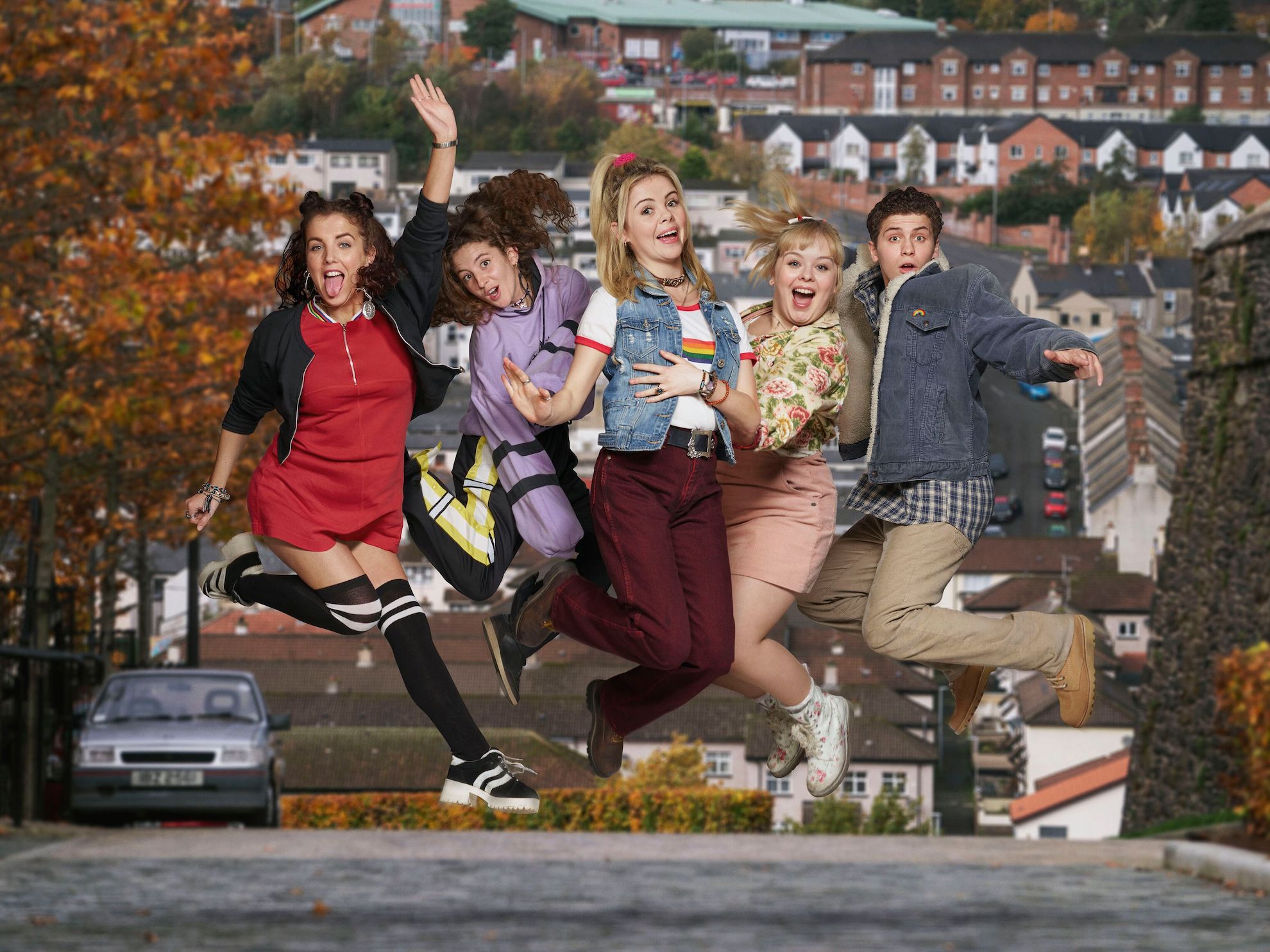 Cast of the Derry Girls jumping while smiling and waving in the middle of a street