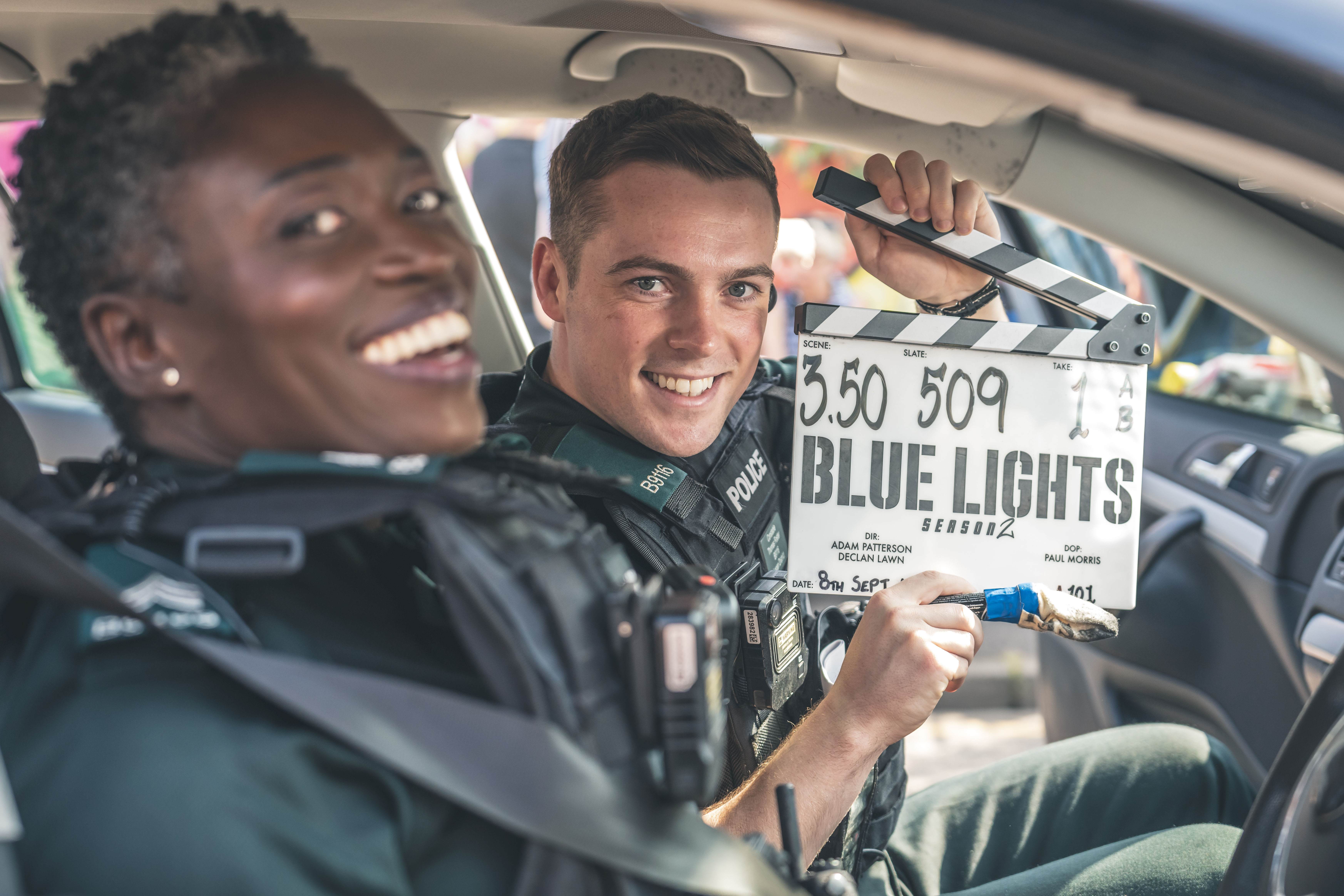 Two actor on the Blue Lights set sit in a police car holding a clapperboard and smiling out of the window