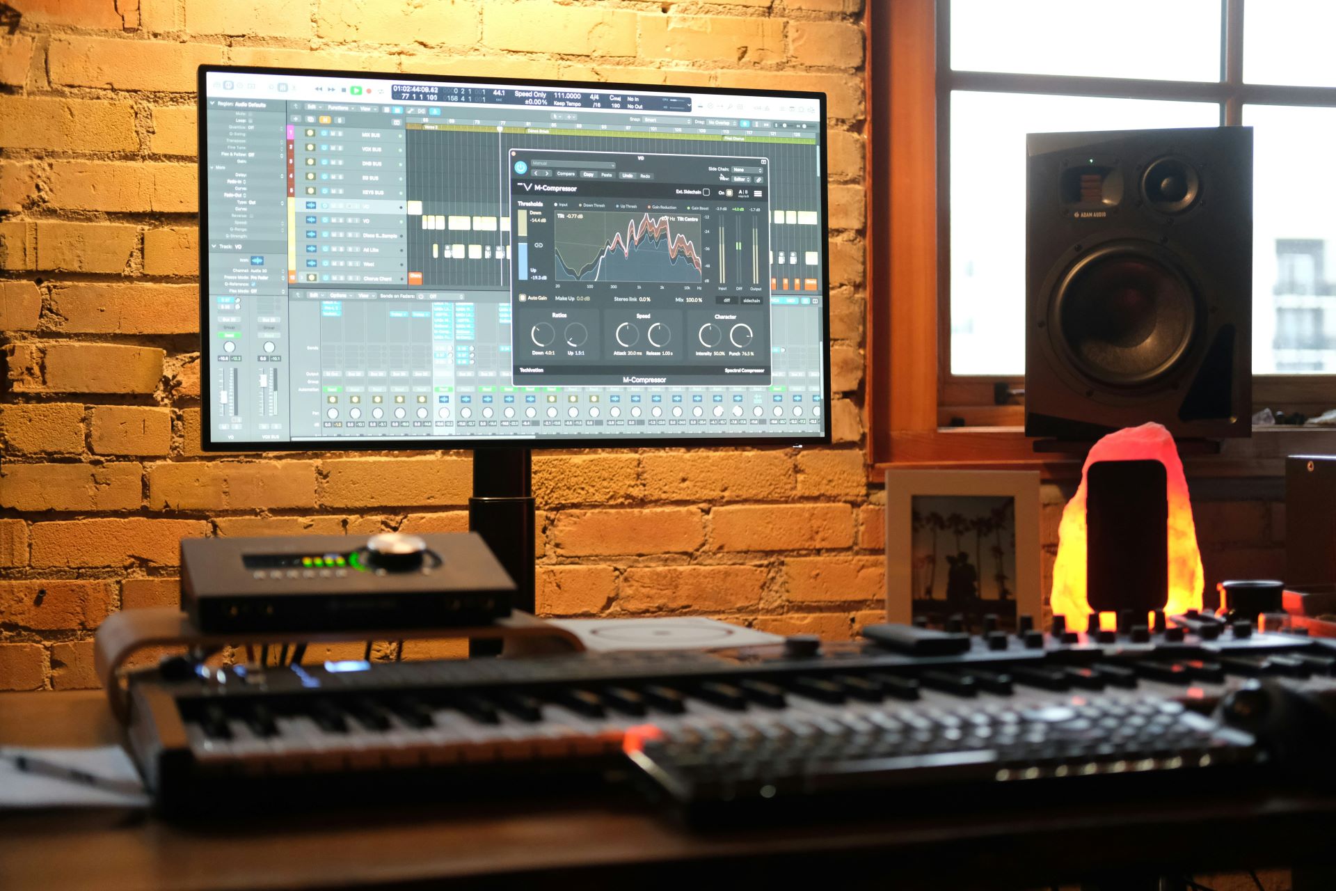 A computer sits on a desk. On the screen, a sound editing software is visible. Beside the desk is a lamp and a speaker.