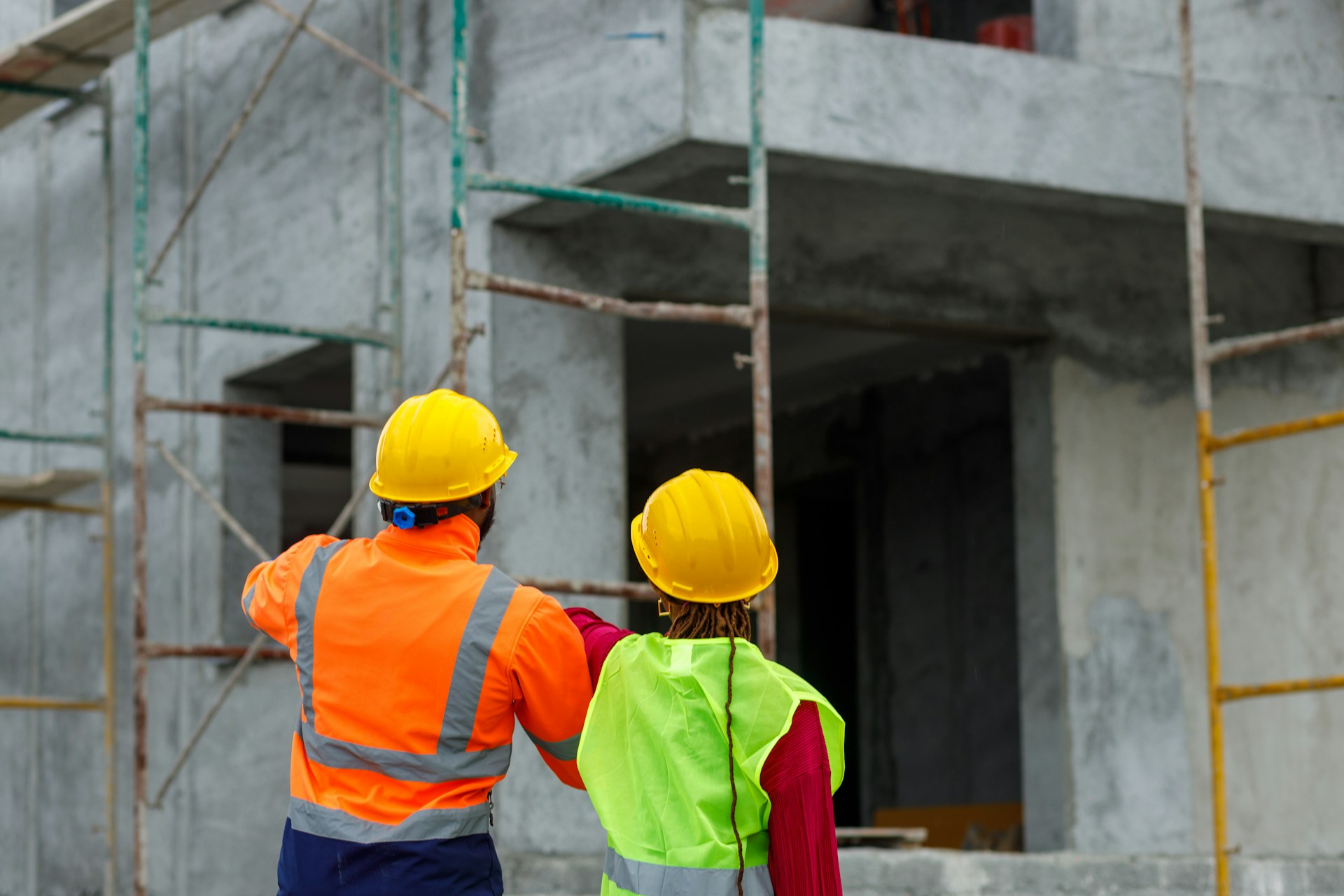 Two people in yellow hard hats and Hi-vis jackets stand looking at a large building under construction