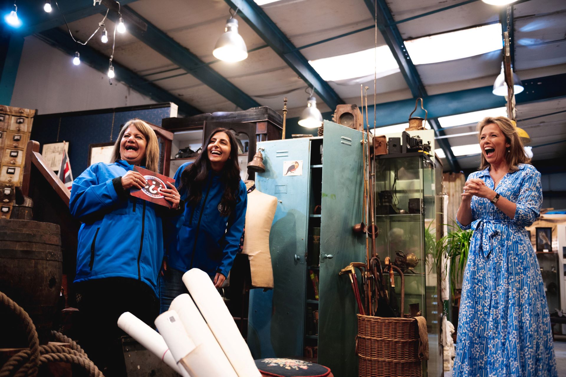Three women stand laughing in a room filled with antiques on the set of Bargain Hunt