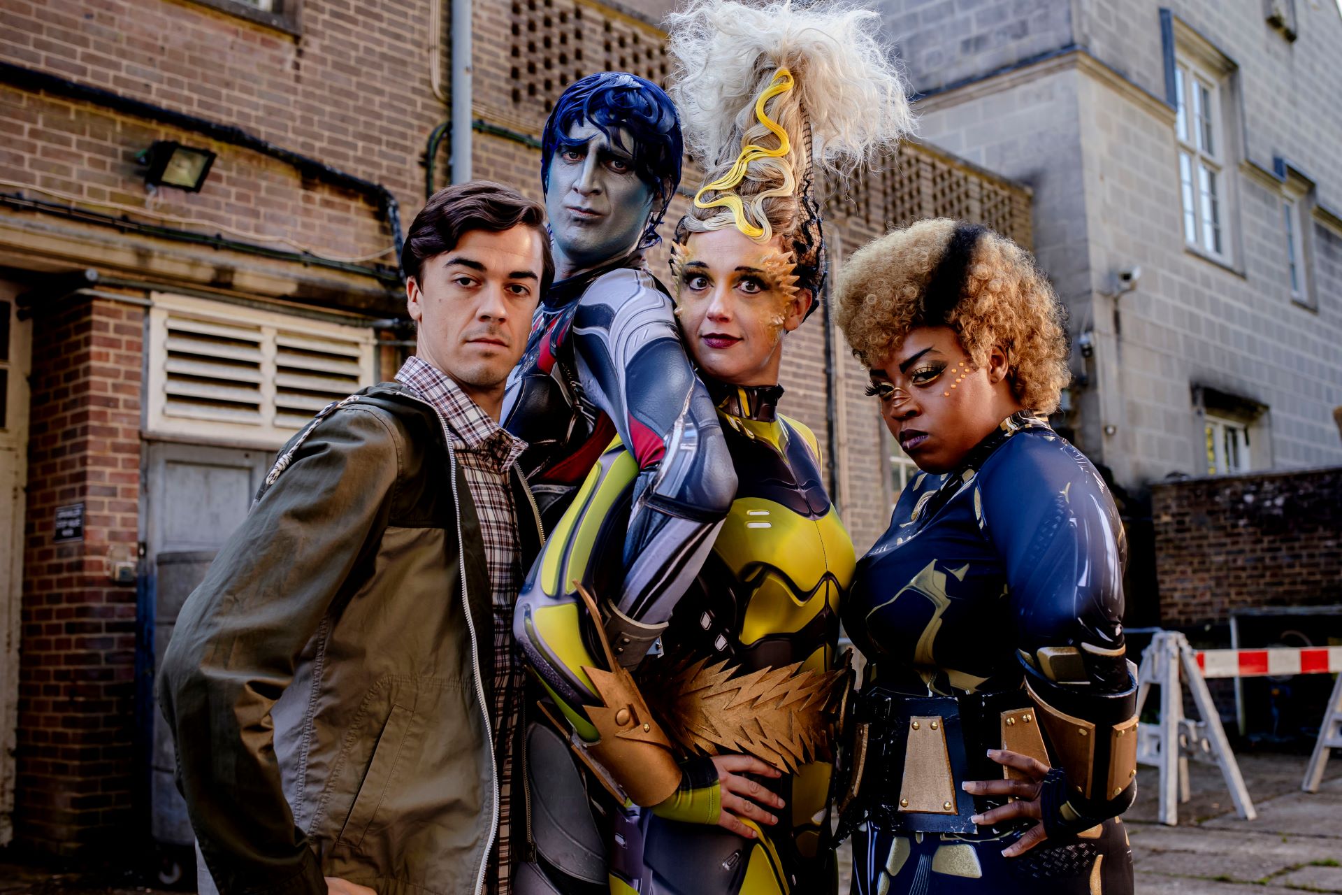 Four people stand posing together, one in casual clothing and three dressed in elaborate superhero costumes