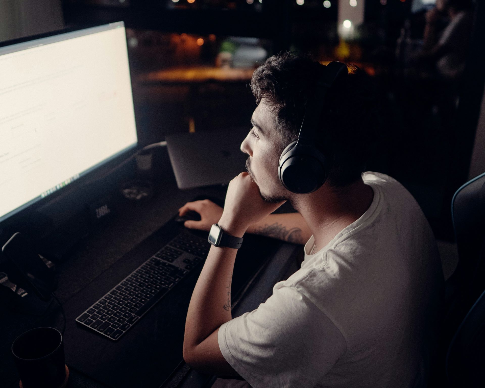 A young man wearing headphones sits in front of a laptop in a dark room in front of a window at night
