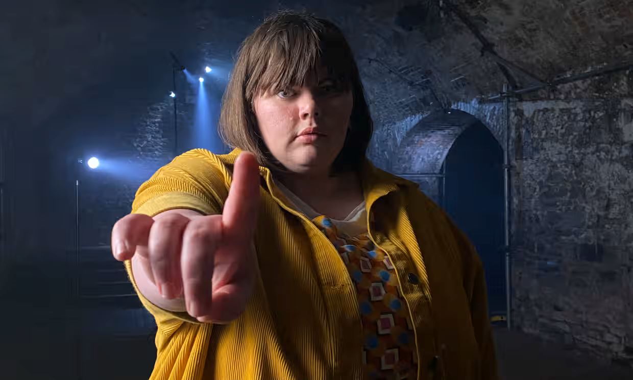 Flo Taylor stands facing the camera with arm held out and her index finger raised