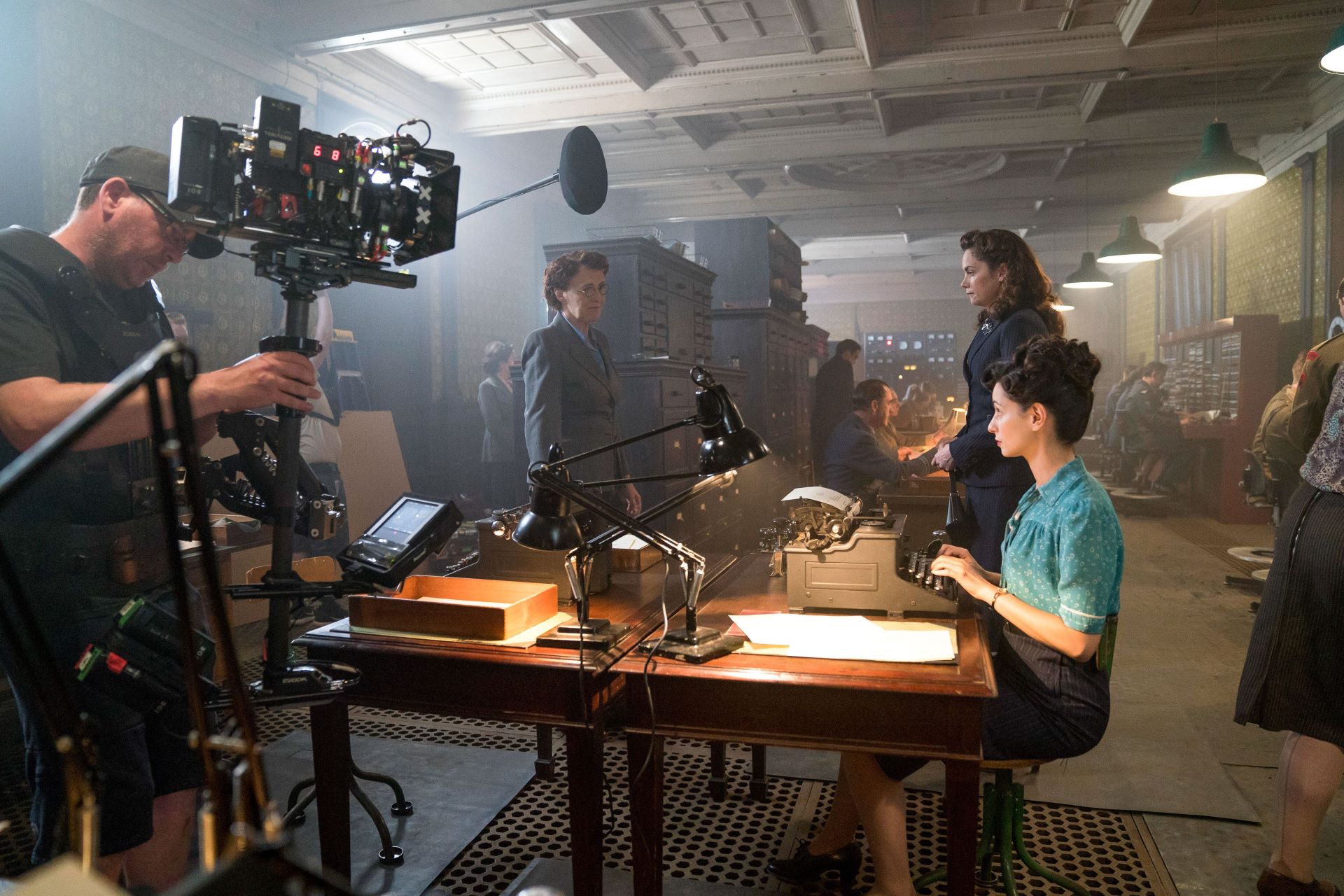 An actress sits at a desk in front of a typewriter on a period drama set. Around her are crewmembers and camera equipment.