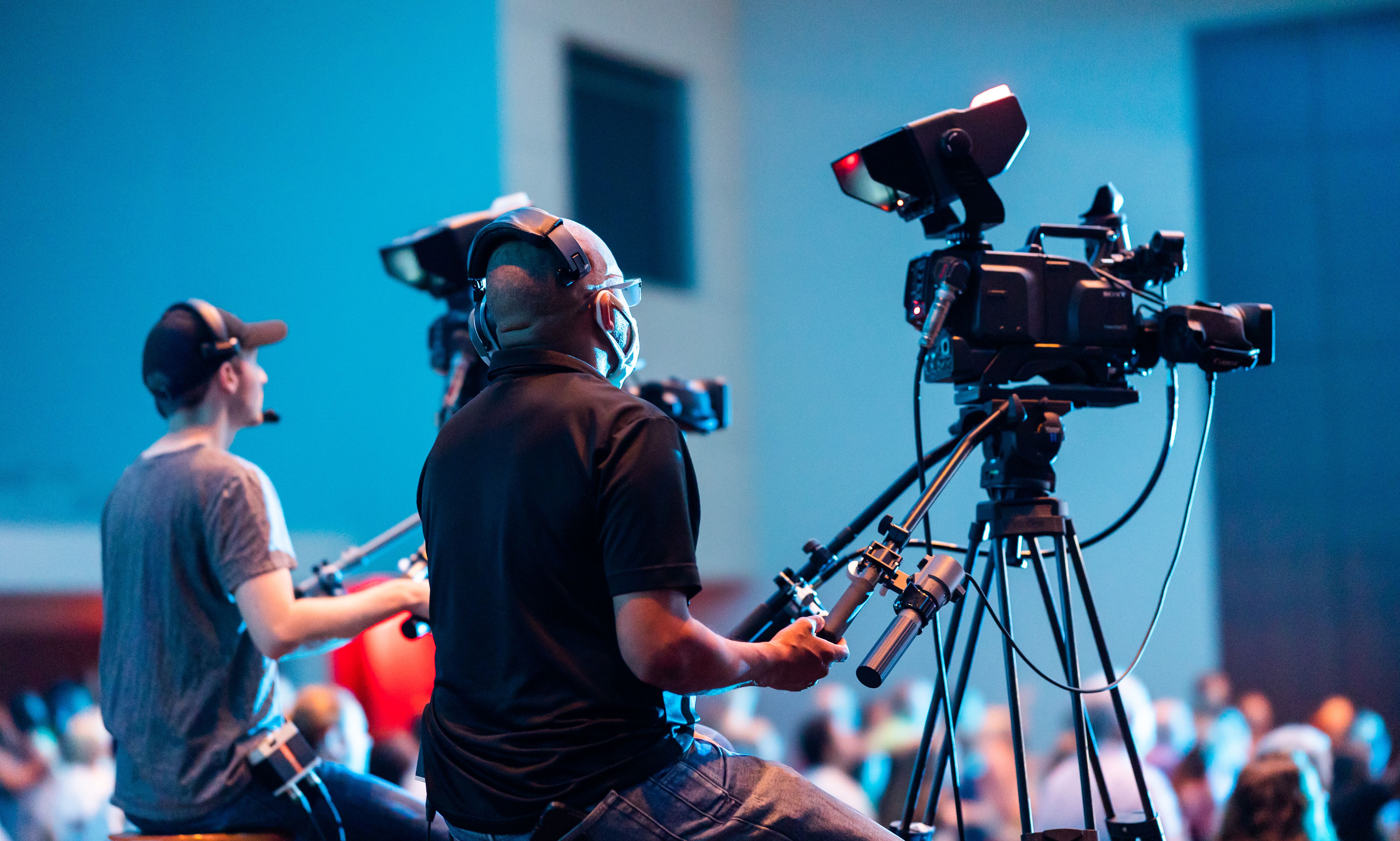 Two camera operators sit managing cameras in a large indoor venue
