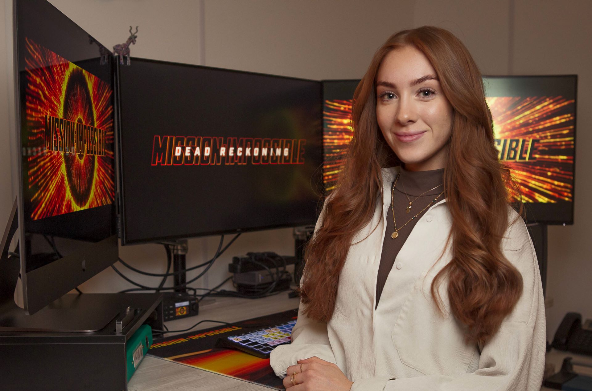 A woman stands smiling towards the camera in front of three monitors displaying the Mission Impossible logo in large bright colours.