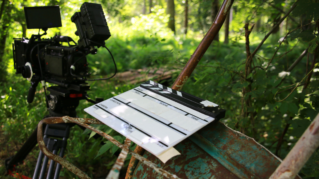 On an outdoor forest production set, a camera sits on the left and a clapperboard rests on an upturned wheelbarrow