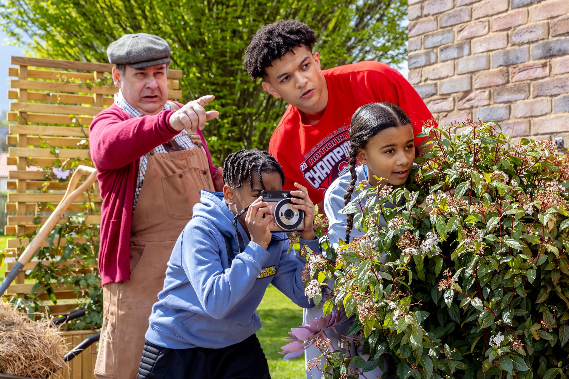 Two men and two children peer around a bush with one child taking a picture on a camera