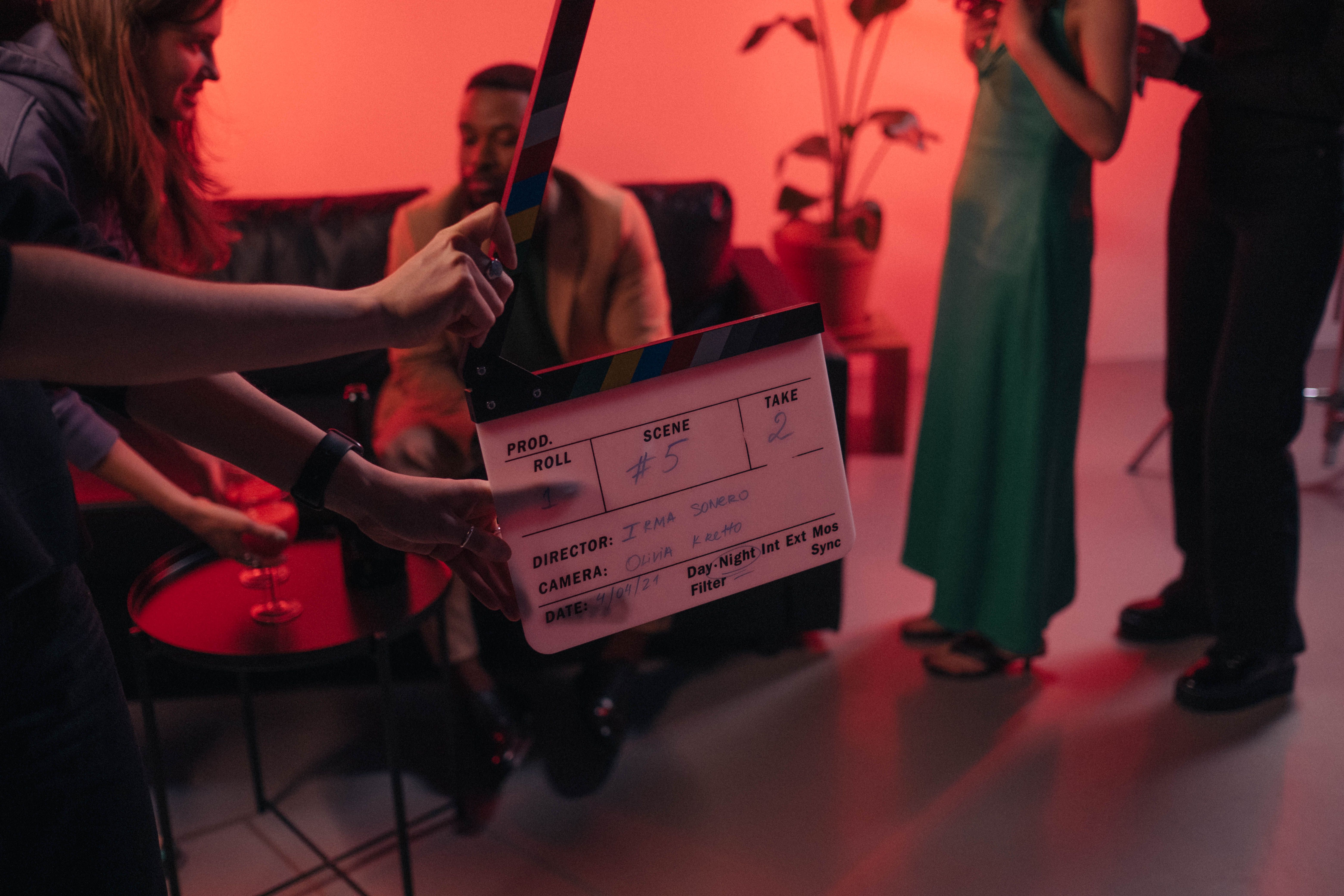 A production set in an indoor setting with low red mood lighting. In the centre someone holds a clapperboard.