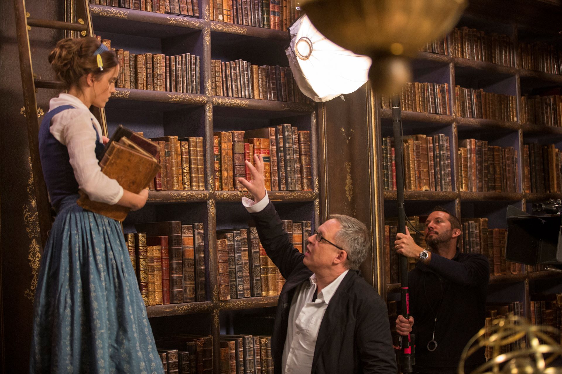 A scene in a library where a director instructs an actress holding some books. In the background, a lights operator adjusts the lighting.